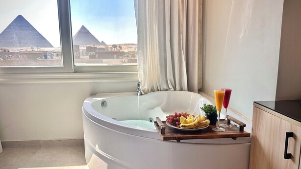 A bathroom with a bathtub, a window with a view of pyramids, and a tray with fruits and drinks. - Tiye Pyramids Hotel (Giza)