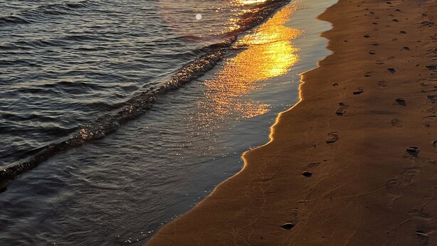 A beach with a sunset over the water, a lighthouse in the distance, and footprints on the sand. - Lakeside Horizon - minutes to Lake Michigan Pere Marquette Beach & Muskegon Lake (Muskegon)