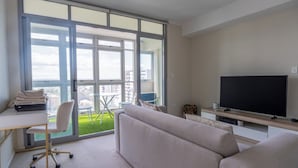 A modern living room with a large window, a flat-screen TV, a sofa, a desk with a chair, and a small balcony with a table and chairs. - Comfy 1BDR Apt 3 minutes to Train Station Burwood (Burwood)