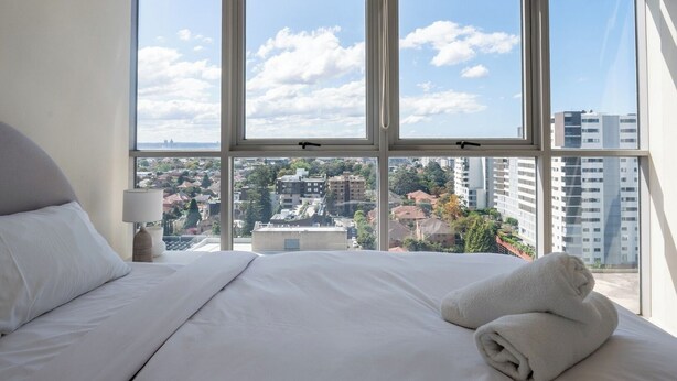 A hotel room with a large bed, white linens, a nightstand with a lamp, and a view of a cityscape through large windows. - Comfy 1BDR Apt 3 minutes to Train Station Burwood (Burwood)