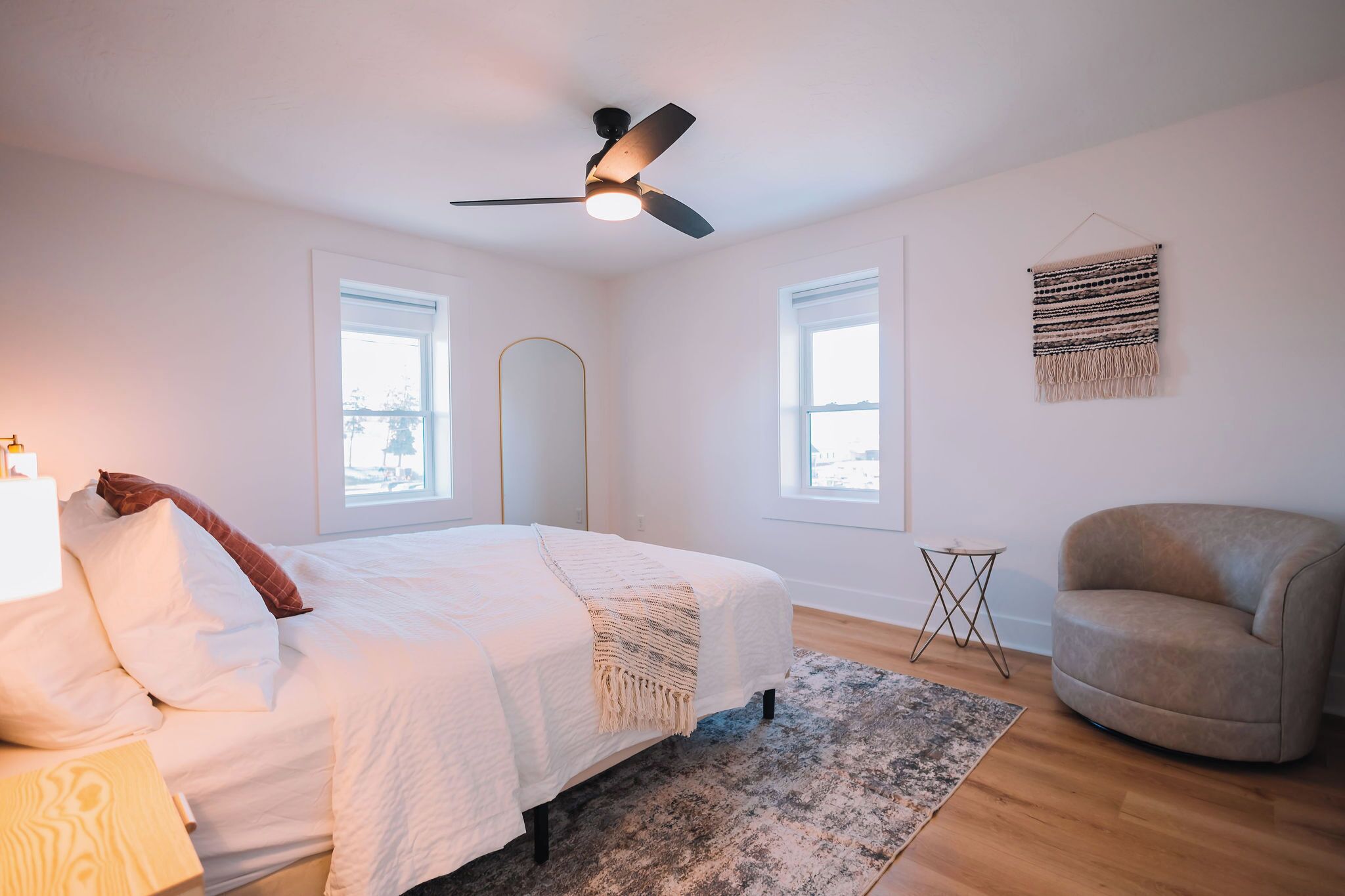 A bedroom with a bed, a chair, a standing lamp, a floor lamp, a ceiling fan, and a wall-mounted tapestry.