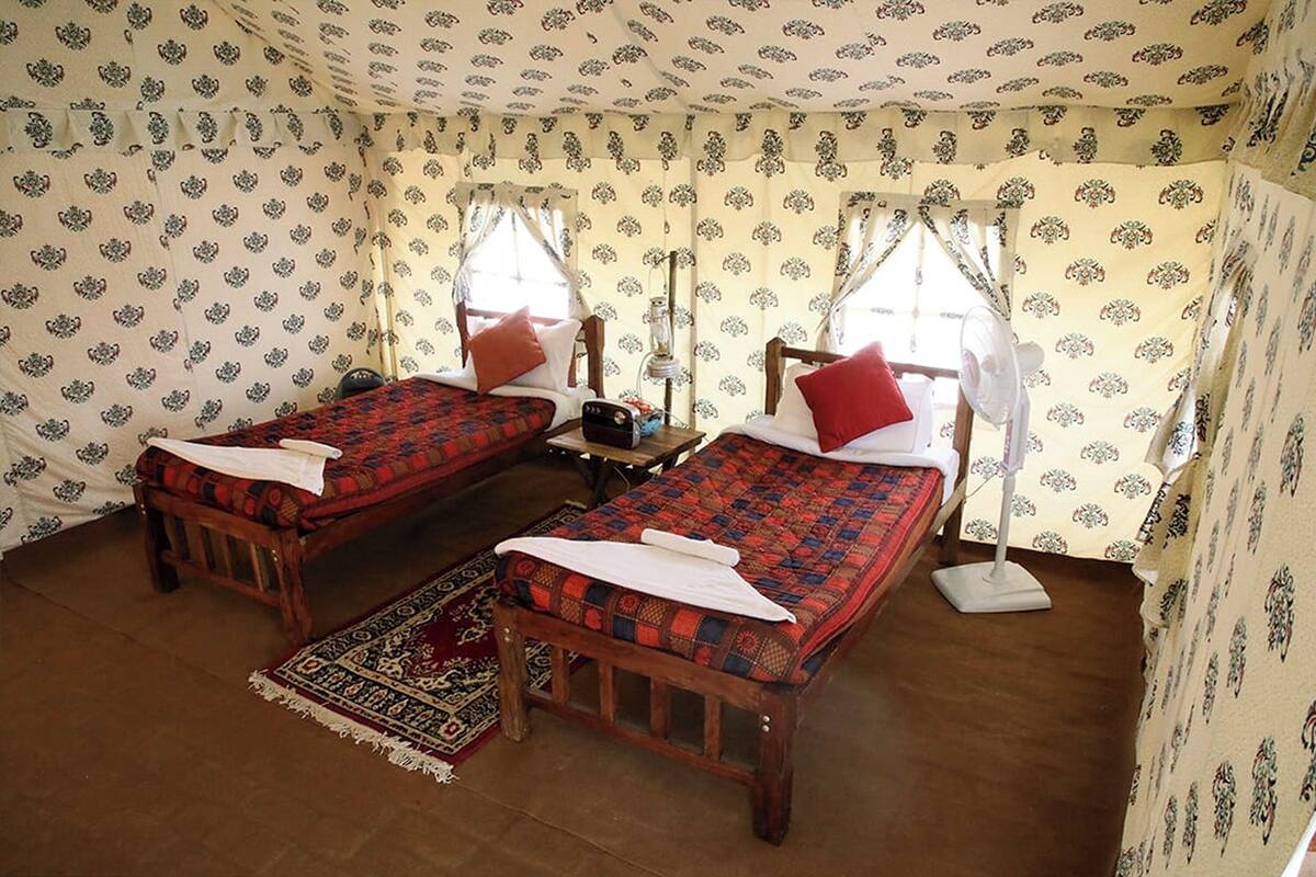Una habitación con dos camas de madera, cada una con colcha roja y azul, almohadas blancas y un banco de madera.