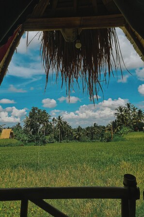 A view from inside a structure with a thatched roof, looking out over a green field with palm trees and a clear sky. - Swaha Retreat & Eco Park Ubud (Sebatu)