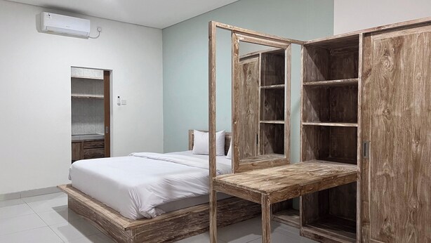 A bedroom with a wooden bed frame, a bedside table, a wardrobe with mirrored doors, and a built-in shelf unit. - Maisaba Living Sibang Gede (Abiansemal)
