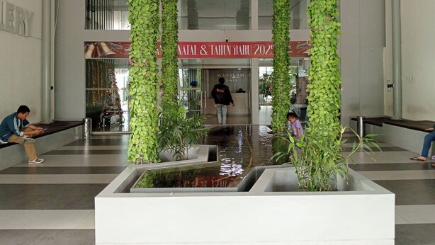 A lobby with a central water feature, vertical greenery, and a reception area with a sign reading "Natal & Tahuin Baru 2025".