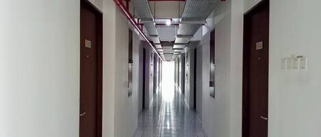 Un pasillo largo y estrecho con puertas a ambos lados, pisos de baldosas y conductos de ventilación en el techo.