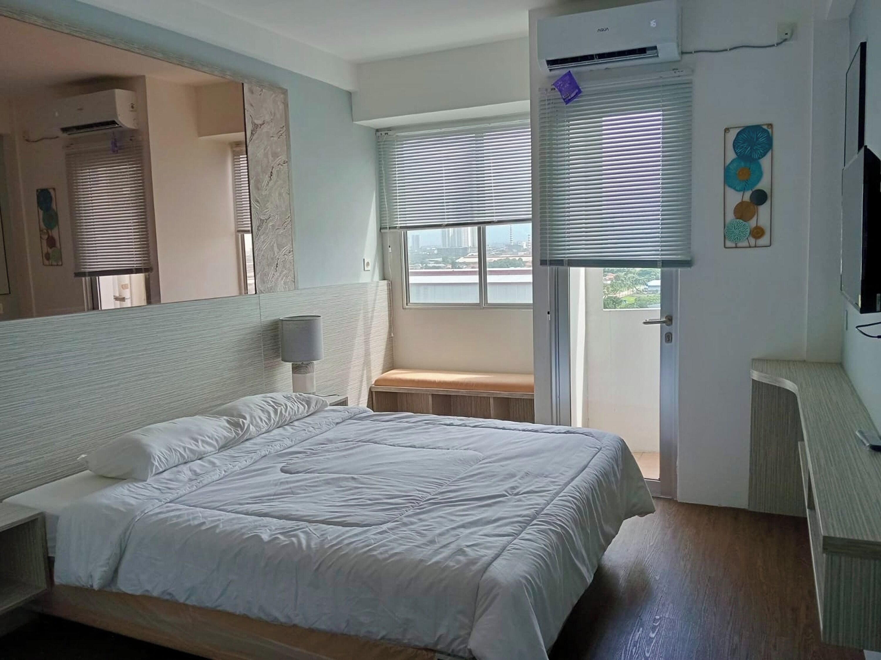 A bedroom with a large bed, a bedside table, a mirror, a window with blinds, and an air conditioning unit.
