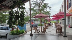 An outdoor cafe area with tables and chairs under umbrellas, surrounded by greenery and a building in the background.