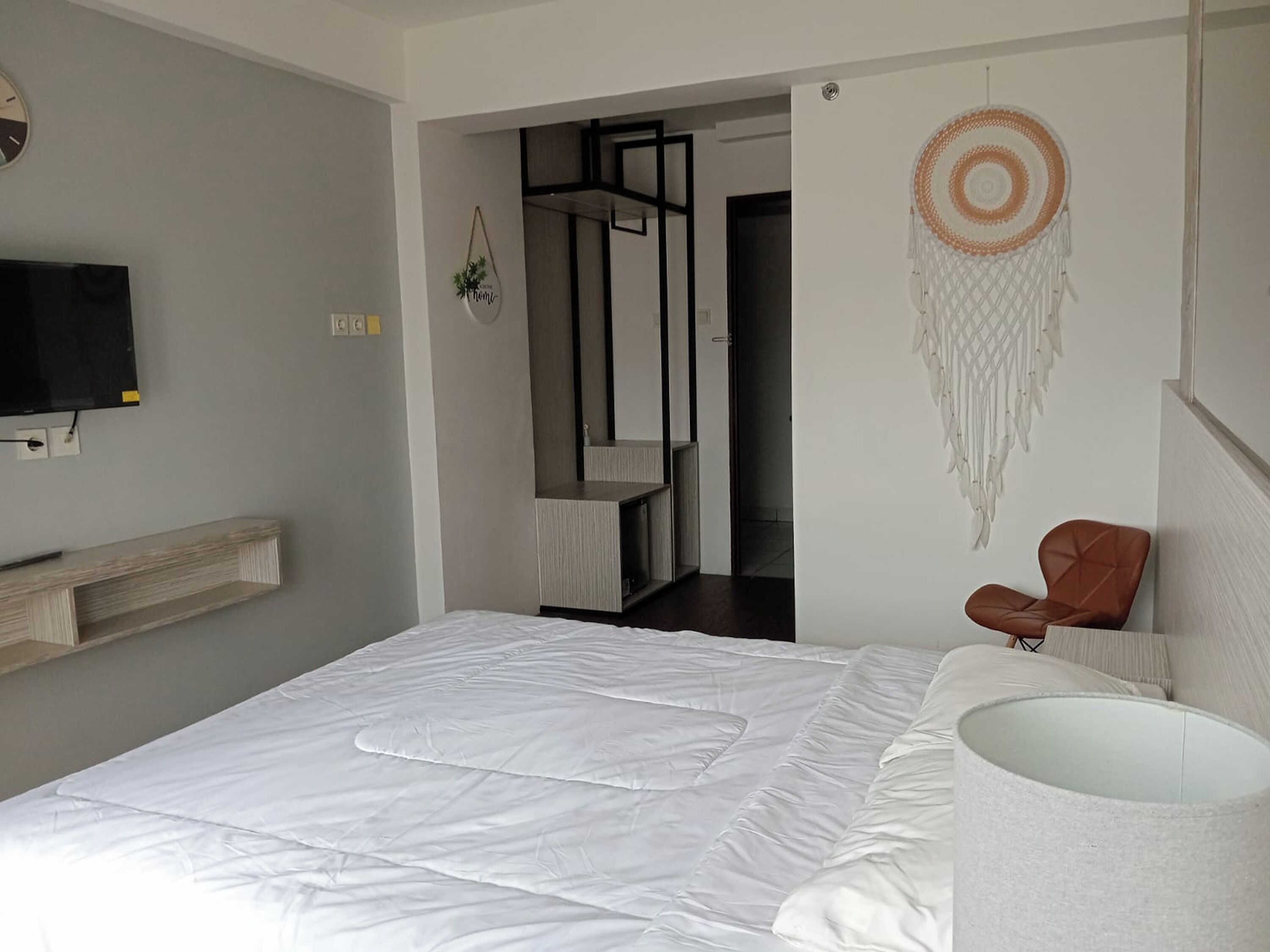 A modern bedroom with a bed, a mounted TV, a wall-mounted shelf, a hanging dreamcatcher, and a chair.