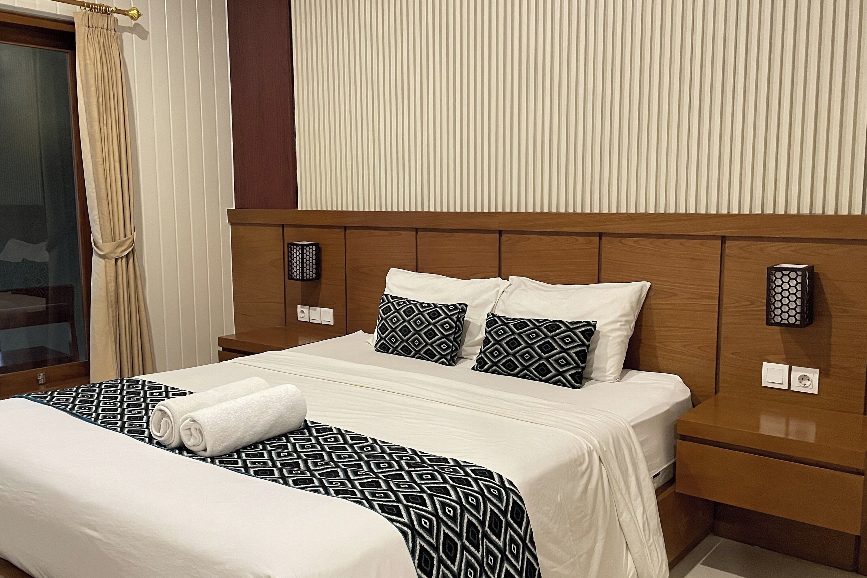 Habitació d'hotel amb un llit gran, dues taules de nit, un panell de capçal de fusta i una finestra amb cortines.
