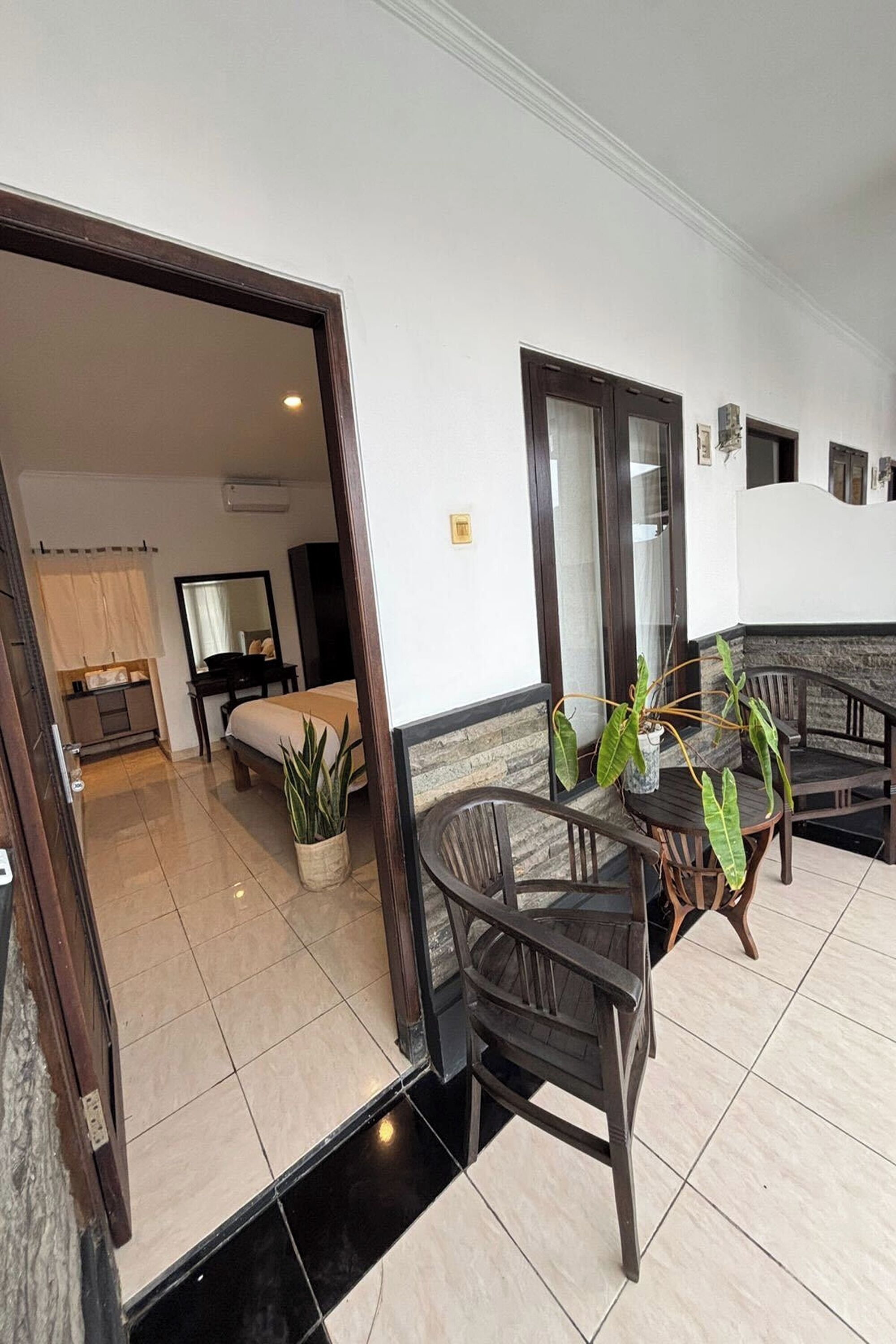 A hotel room with a bed, a small bedside table, a mirror, and a potted plant. There is a dining area with a table and chairs, and a view of the outside.