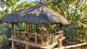 A thatched-roof pavilion with a wooden deck, surrounded by lush greenery and a bamboo fence. - Surfer'S Villa (San Juan)
