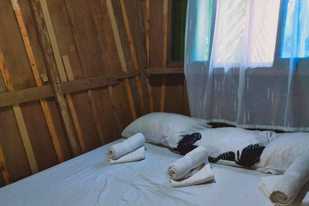 A single bed with white linens and rolled towels, wooden walls, and a window with sheer curtains.