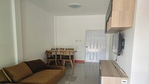 Un salon avec un canapé marron, une table à manger en bois entourée de chaises, un téléviseur fixé au mur et une porte visible au fond.