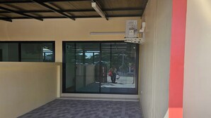 Un espace extérieur abrité, doté d’un sol carrelé, d’une porte d’entrée en verre et d’une boîte électrique visible sur le mur.