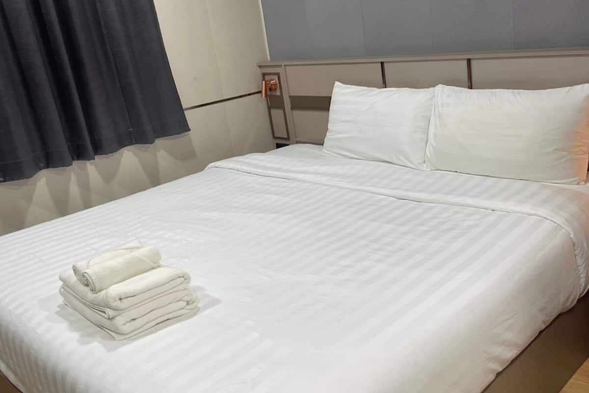 A neatly made bed with white linens and pillows, a folded white towel on the footboard, and a dark curtain on the left.