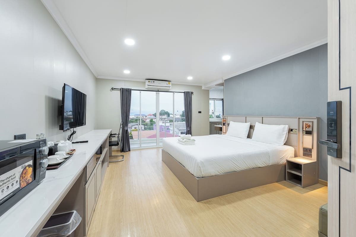 A modern hotel room with a large bed, a flat-screen TV, a kitchenette, and a view of the city through large windows.