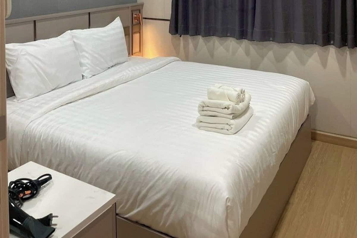 A hotel room with a large bed, a bedside table with a phone and charger, a window with dark curtains, and a wall-mounted light.