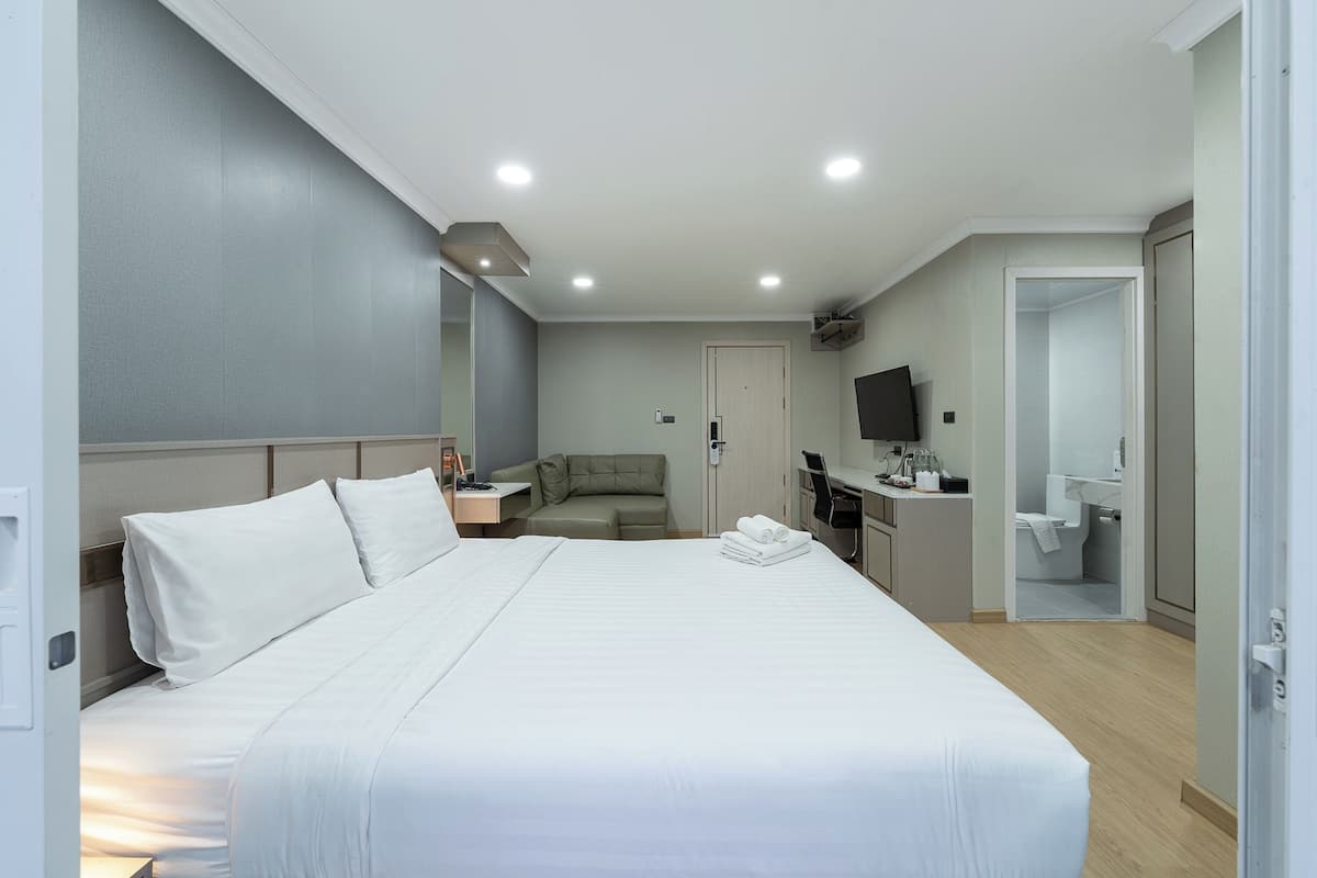 A hotel room with a large bed, a desk, a sofa, a TV, and a bathroom visible through an open door.