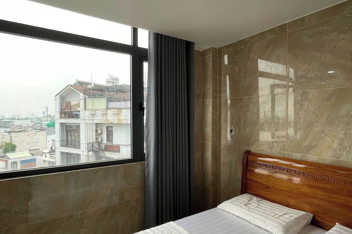 Ein Hotelzimmer mit einem großen Bett, einem Holzkopfende, Blick auf Gebäude durch ein Fenster und einem Vorhang.