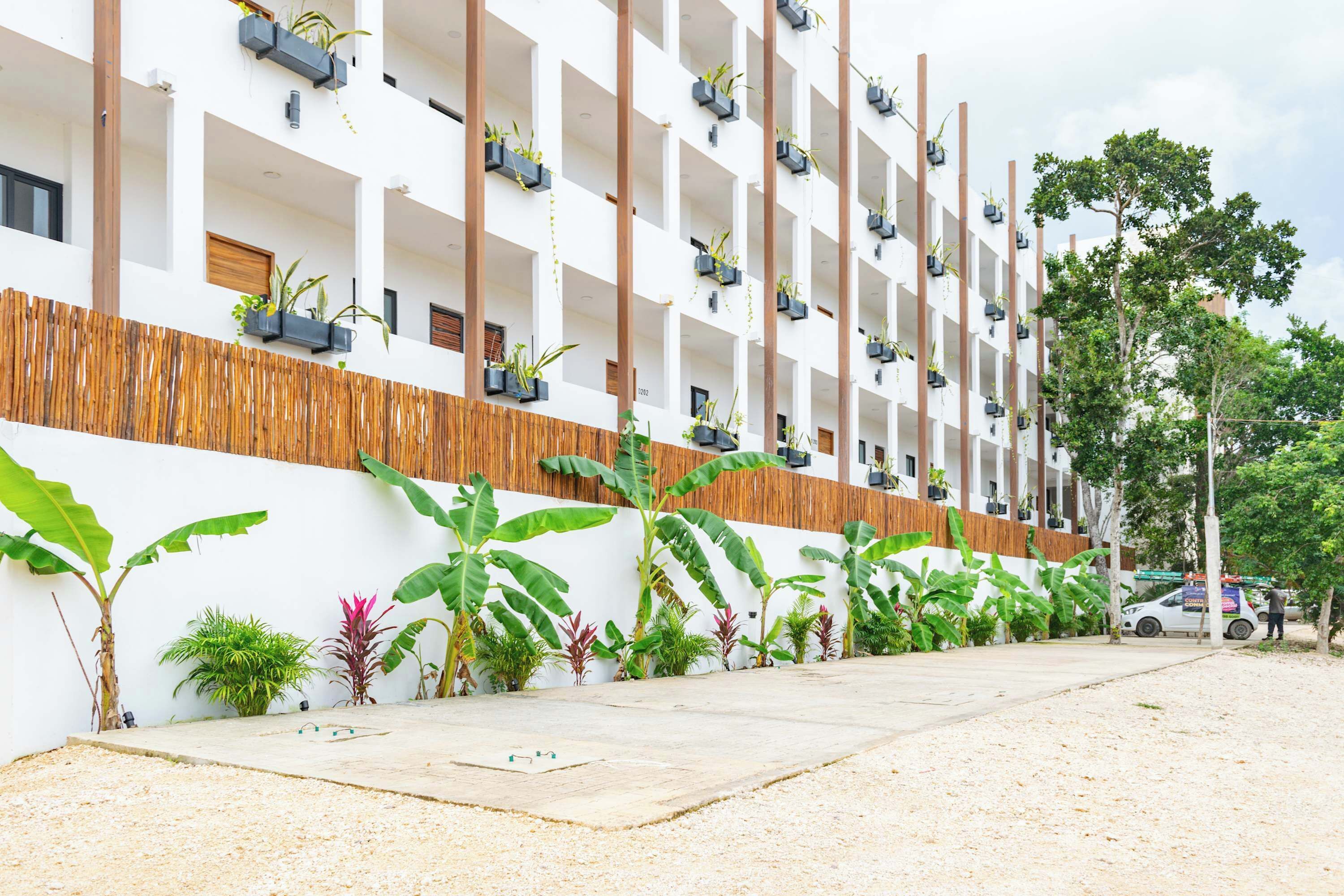 Un edificio moderno con varios balcones, cada uno adornado con plantas en macetas. Se aprecia una cerca de madera y diversas plantas tropicales a lo largo de la pared.