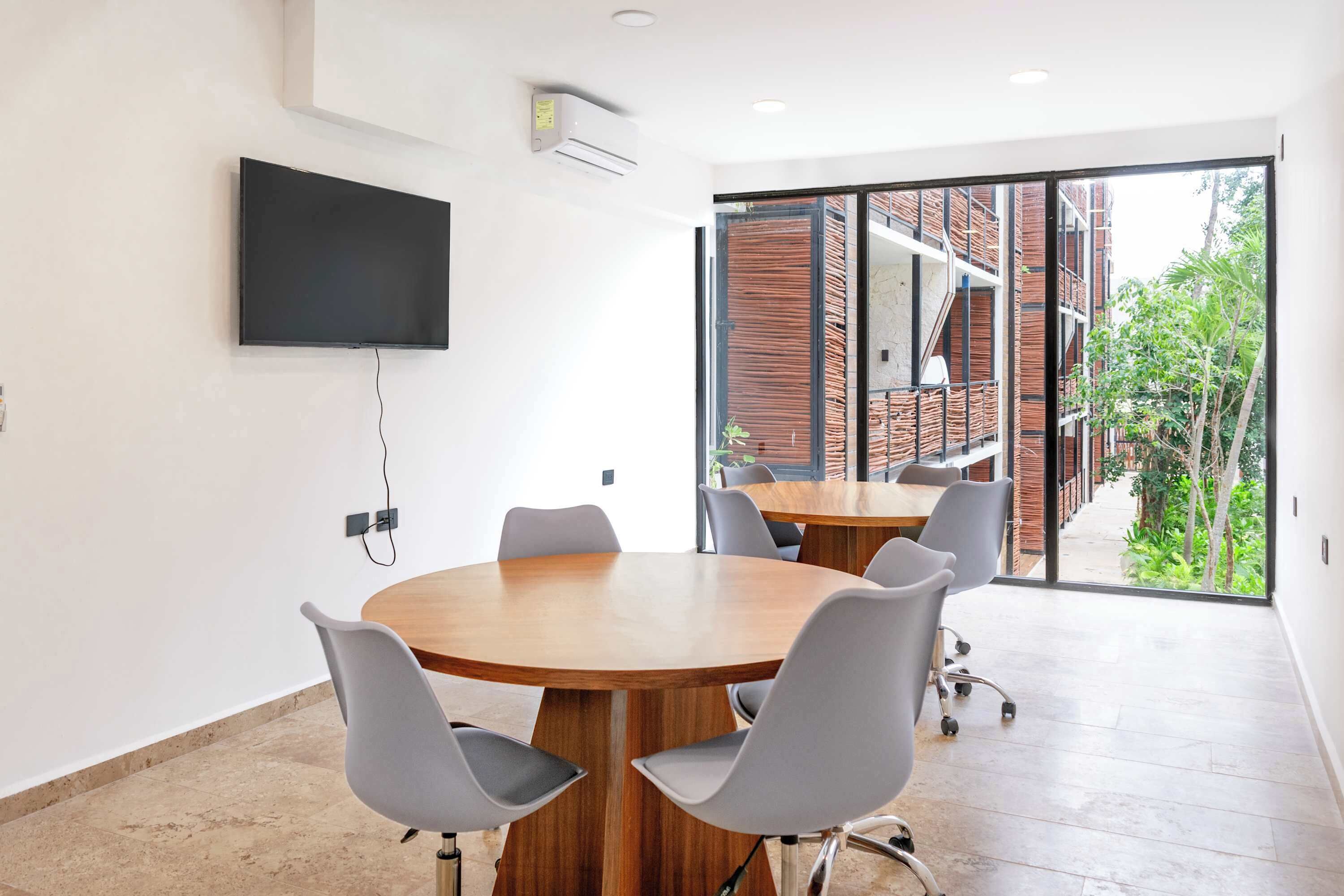 A conference room with a large table, six chairs, a flat-screen TV mounted on the wall, and a large window offering a view of a brick building and greenery outside.