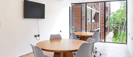 A conference room with a large table, six chairs, a flat-screen TV mounted on the wall, and a large window offering a view of a brick building and greenery outside.