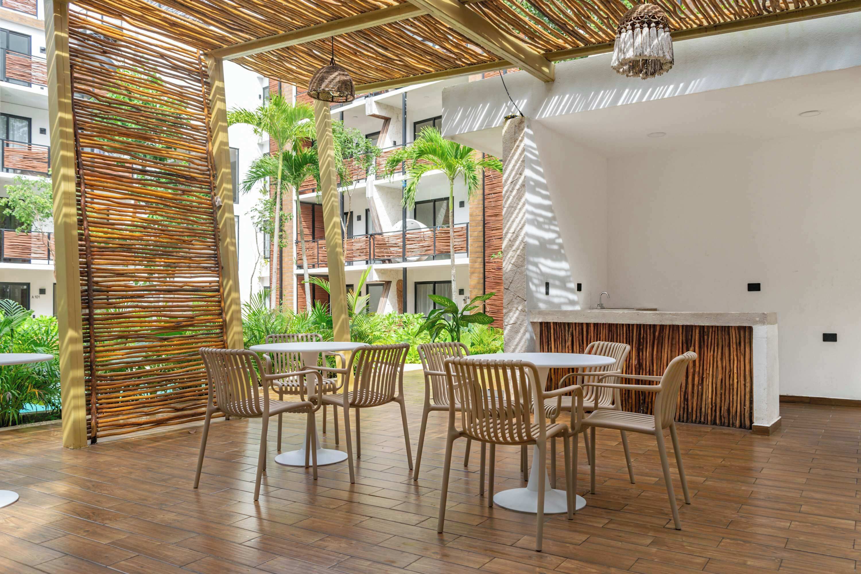 A modern outdoor patio with wooden chairs and tables, surrounded by a bamboo fence, and overlooking a pool and greenery.
