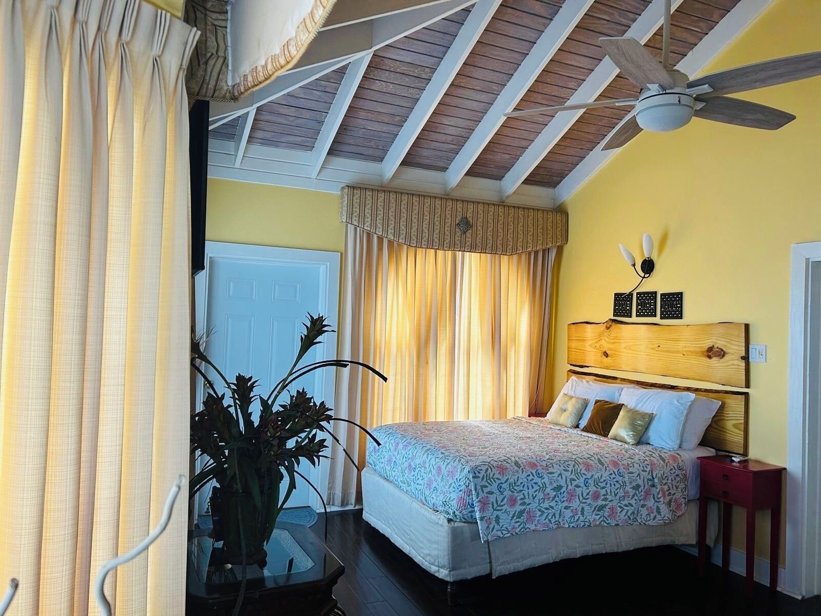 Un dormitorio con una cama, un ventilador de techo, una mesita de noche y una planta en maceta.