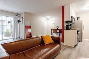 A modern living room with a brown leather sofa, a dining area with a red accent wall, and a kitchen with a stainless steel refrigerator and coffee maker.