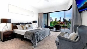 A modern bedroom with a city view, a bed with a grey duvet, a nightstand, a chair, and a TV on the wall.