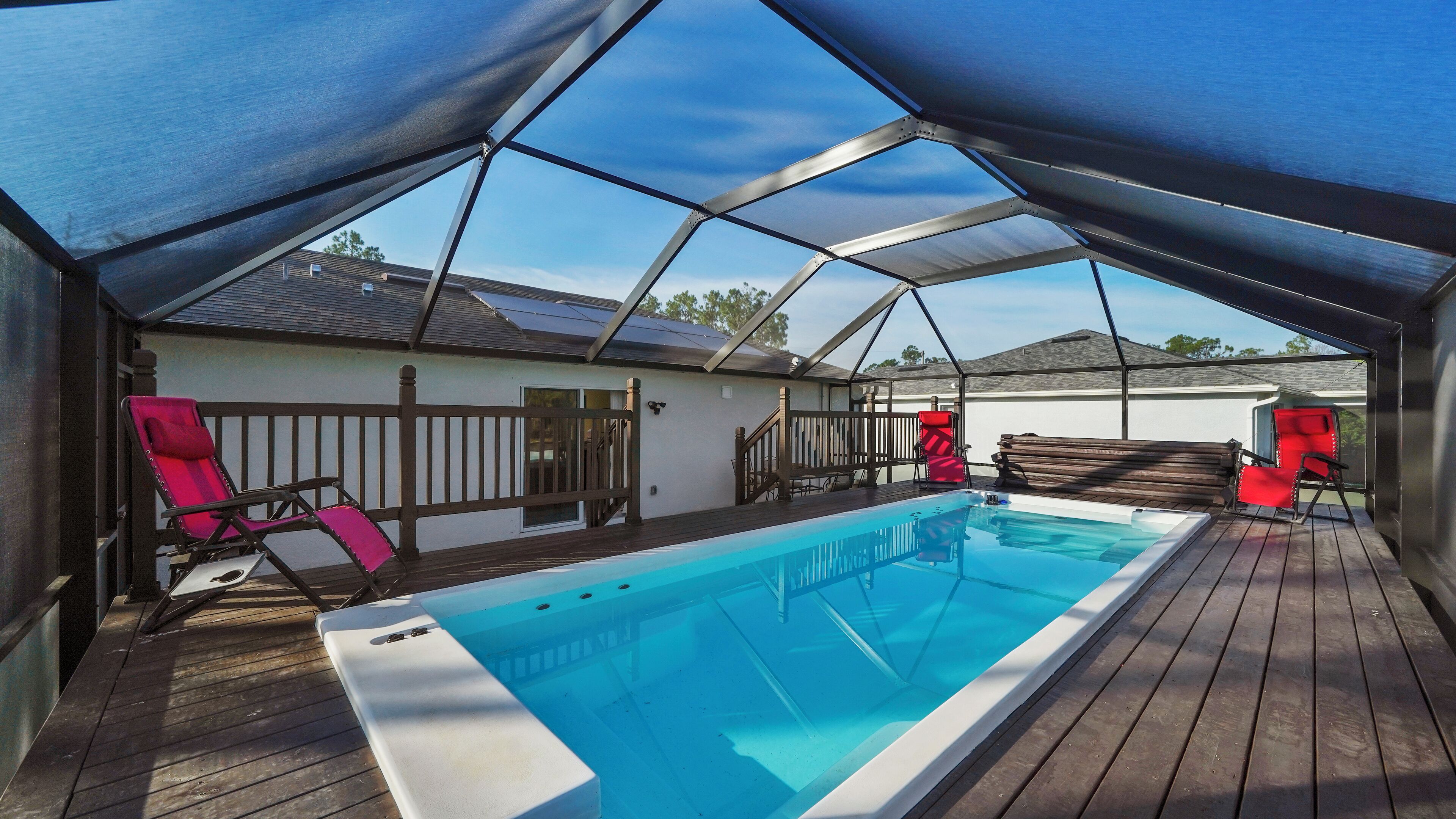 A covered outdoor swimming pool with wooden decking, a white pool edge, and two red lounge chairs.