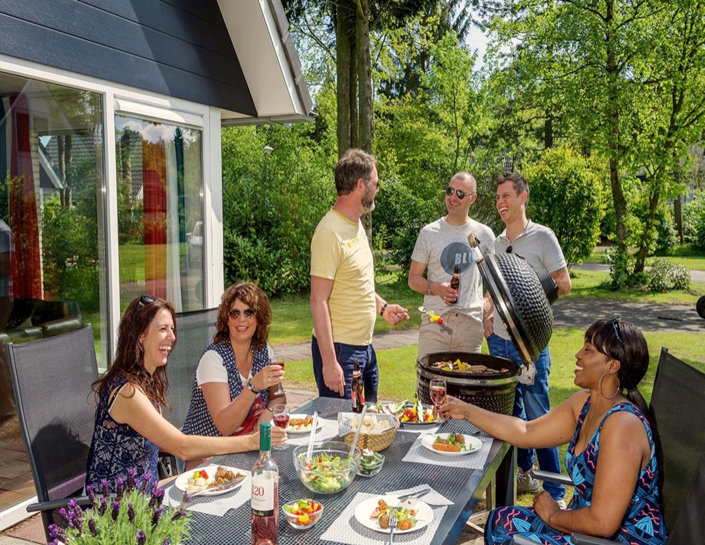 An outdoor dining area with a table set for a meal, surrounded by greenery and a building with large windows.