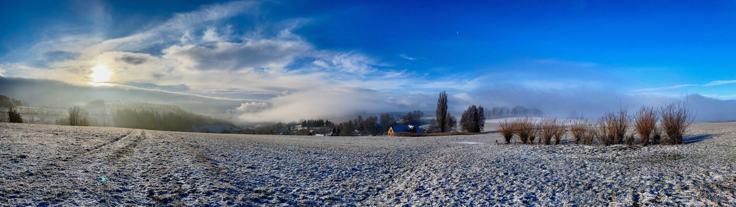 A panoramic view of a snow-covered landscape with a clear sky, a distant sun, and a few buildings in the background.