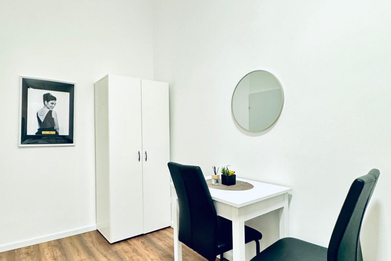 Una habitación con un armario blanco, una mesita con dos sillas, un espejo redondo y un cuadro enmarcado en la pared.