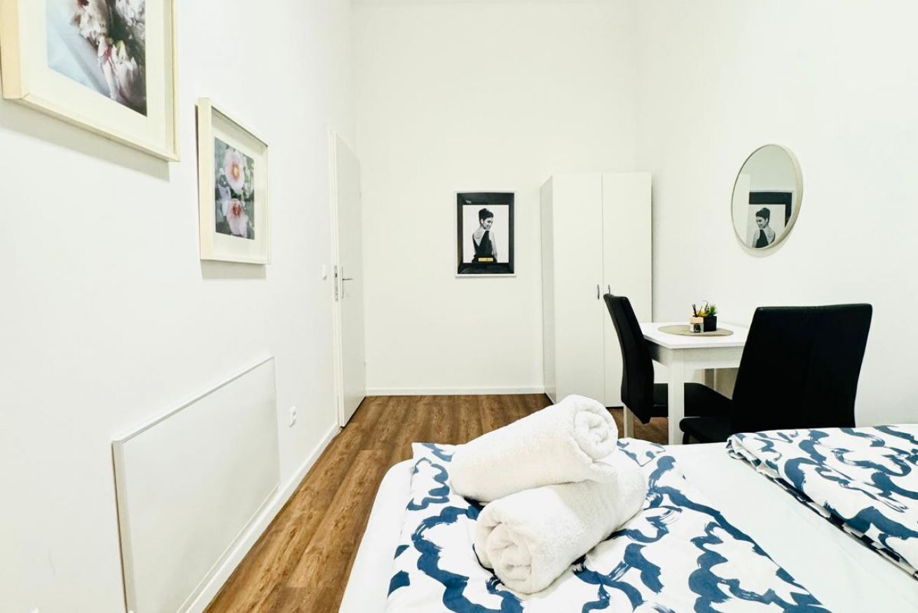 A hotel room with a bed, a desk, a chair, a mirror, and framed pictures on the wall.