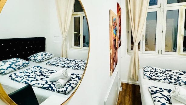 Una habitación de hotel con un espejo redondo, dos camas con ropa de cama azul y blanca estampada, un cabecero de madera y una ventana con cortinas.