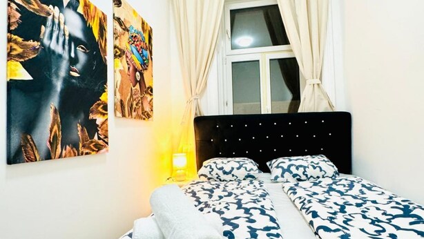 A bedroom with a bed, two paintings, a window with curtains, and a nightstand with a lamp.