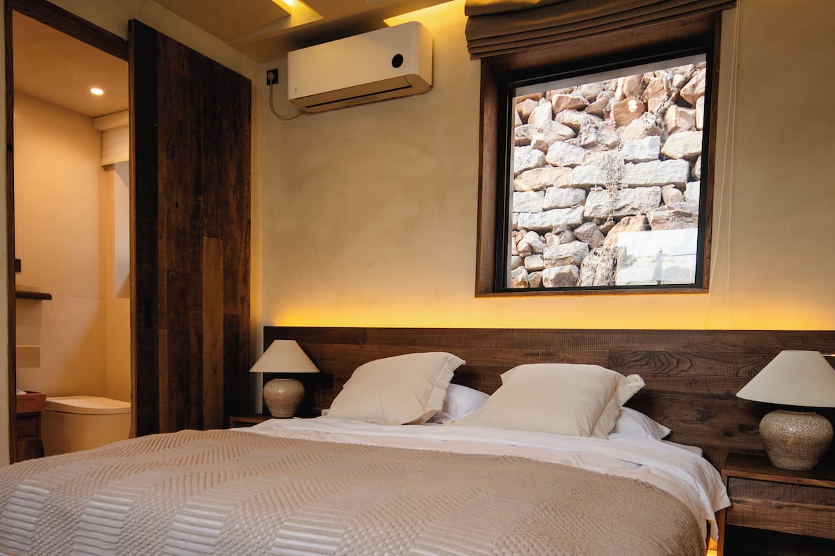 Un dormitorio con una cama grande, cabecera de madera, dos lámparas de noche y vista a una pared de piedra.