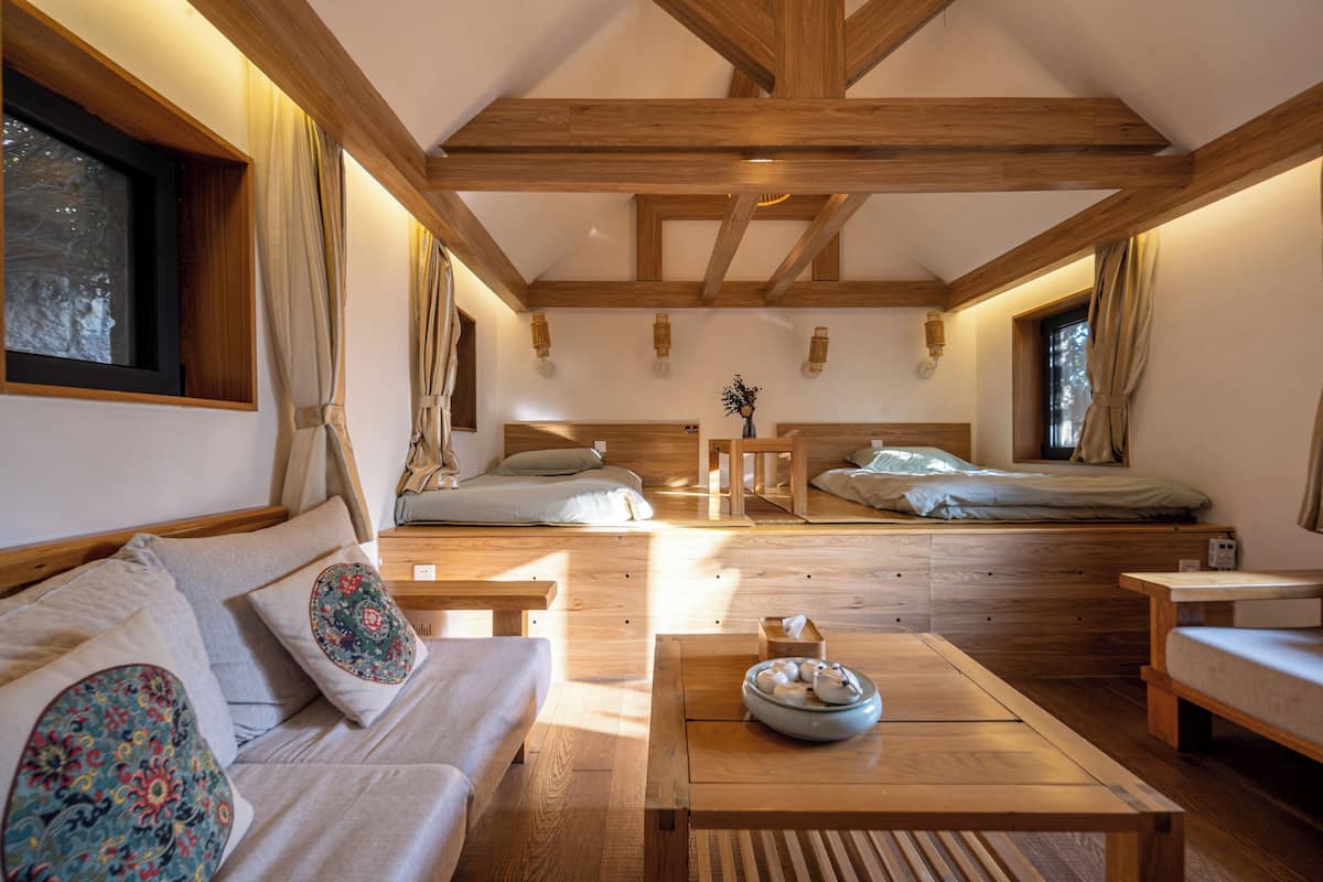 Una habitación acogedora y luminosa con vigas de madera, un sofá con cojines estampados, una mesita con un cuenco y una cama con cabecero.