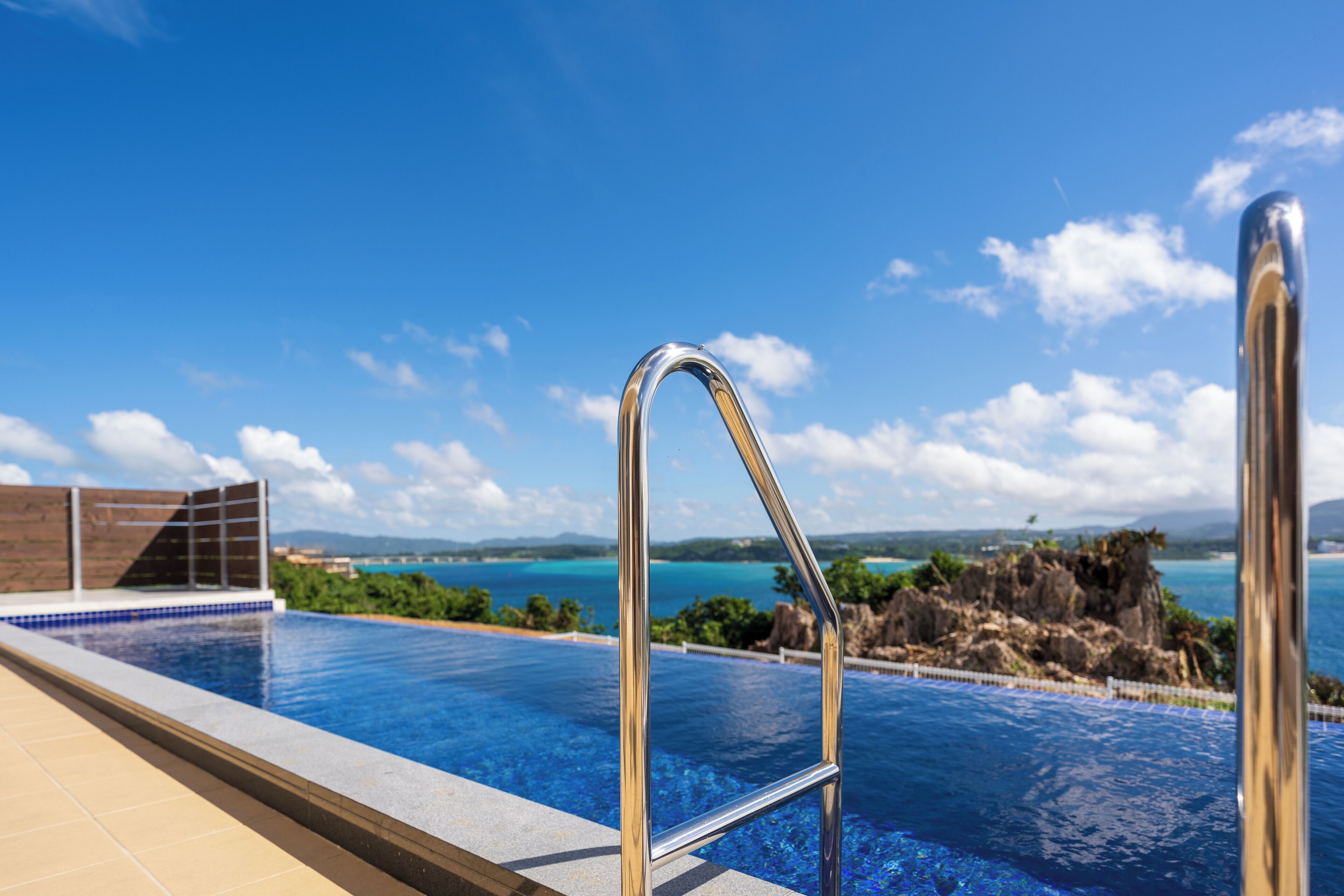 Una piscina con escalera metálica, solárium con baldosas y vista al océano y paisaje rocoso.