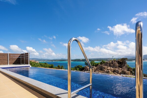 A swimming pool with a metal ladder, a tiled deck, and a view of the ocean and rocky landscape. - Pool Villa Kouri The Sweet (Nakijin)