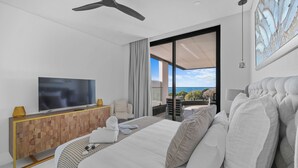 Ein modernes Hotelzimmer mit einem großen Bett, einem Flachbildfernseher, einer Holztruhe, einem Balkon mit Blick und einem Deckenventilator.