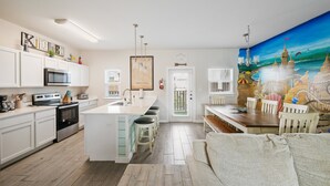 Een moderne keuken met witte kasten, een centraal eiland met krukken, een eethoek met een houten tafel en stoelen, en een muurschildering van een zeegezicht.