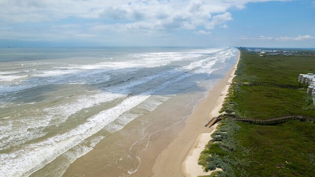 Luchtbeeld van een strand met golven, een grasveld en gebouwen in de verte.