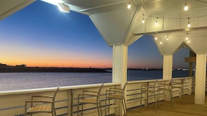 Een houten terras met stoelen en een reling, uitkijkend over een wateroppervlak bij zonsondergang.