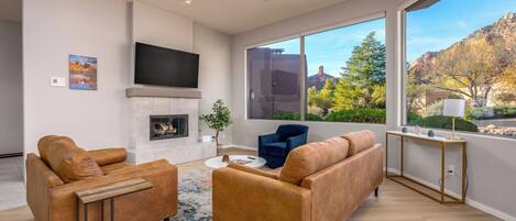 A modern living room with a fireplace, leather sofas, a coffee table, and a large window offering a view of trees and mountains.
