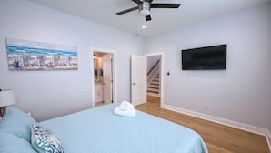 A bedroom with a bed, a ceiling fan, a painting of a beach scene, a TV mounted on the wall, and a staircase leading to another room. - Modern Lux Home 2 Mins to the Beach+Putting Green
 (Galveston)