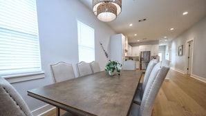 A dining area with a wooden table and upholstered chairs, a potted plant, and a kitchen in the background. - Modern Lux Home 2 Mins to the Beach+Putting Green
 (Galveston)