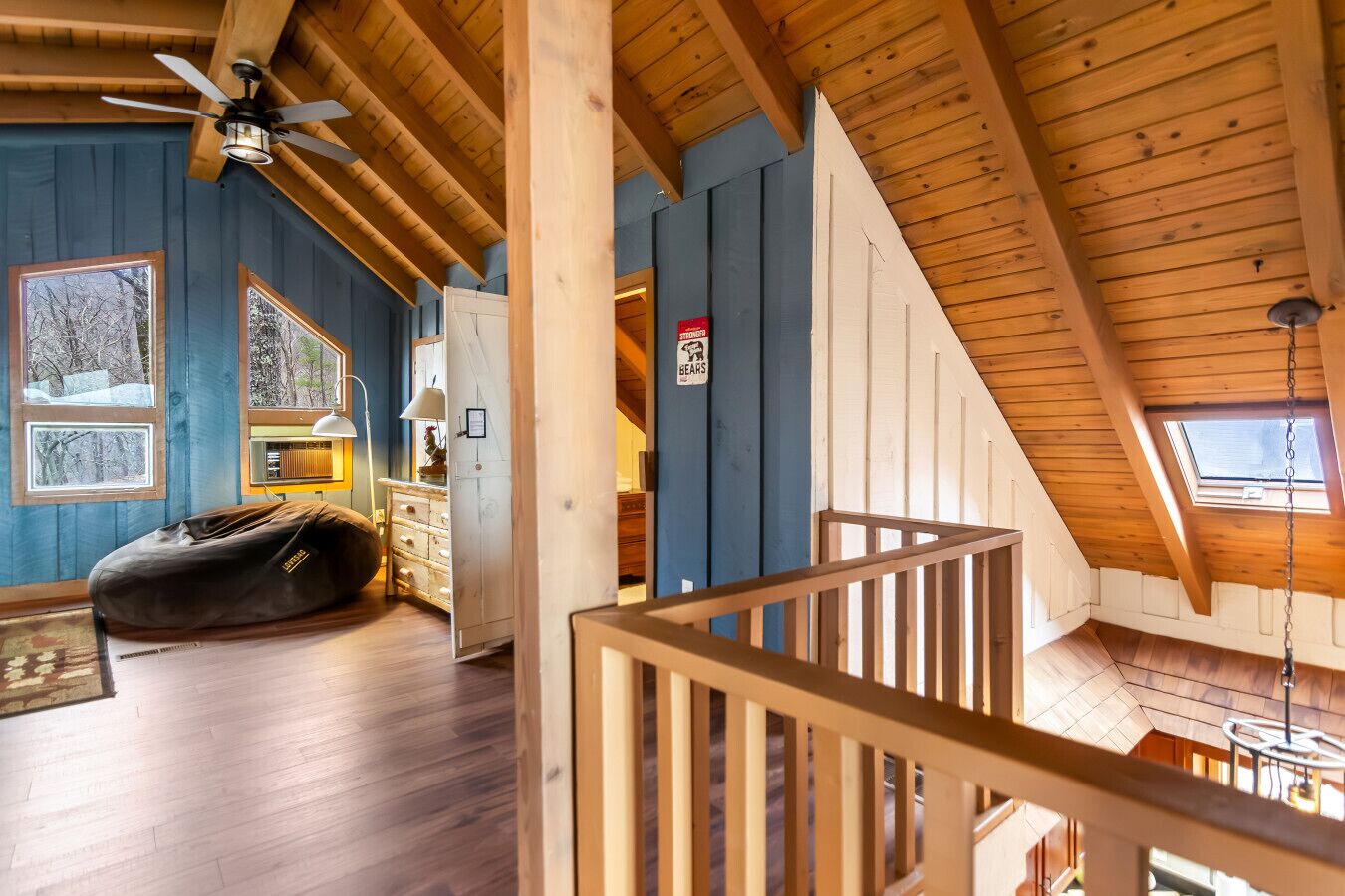 A cozy cabin interior with a loft bedroom, wooden beams, a ceiling fan, a bean bag, and a view of the outdoors.